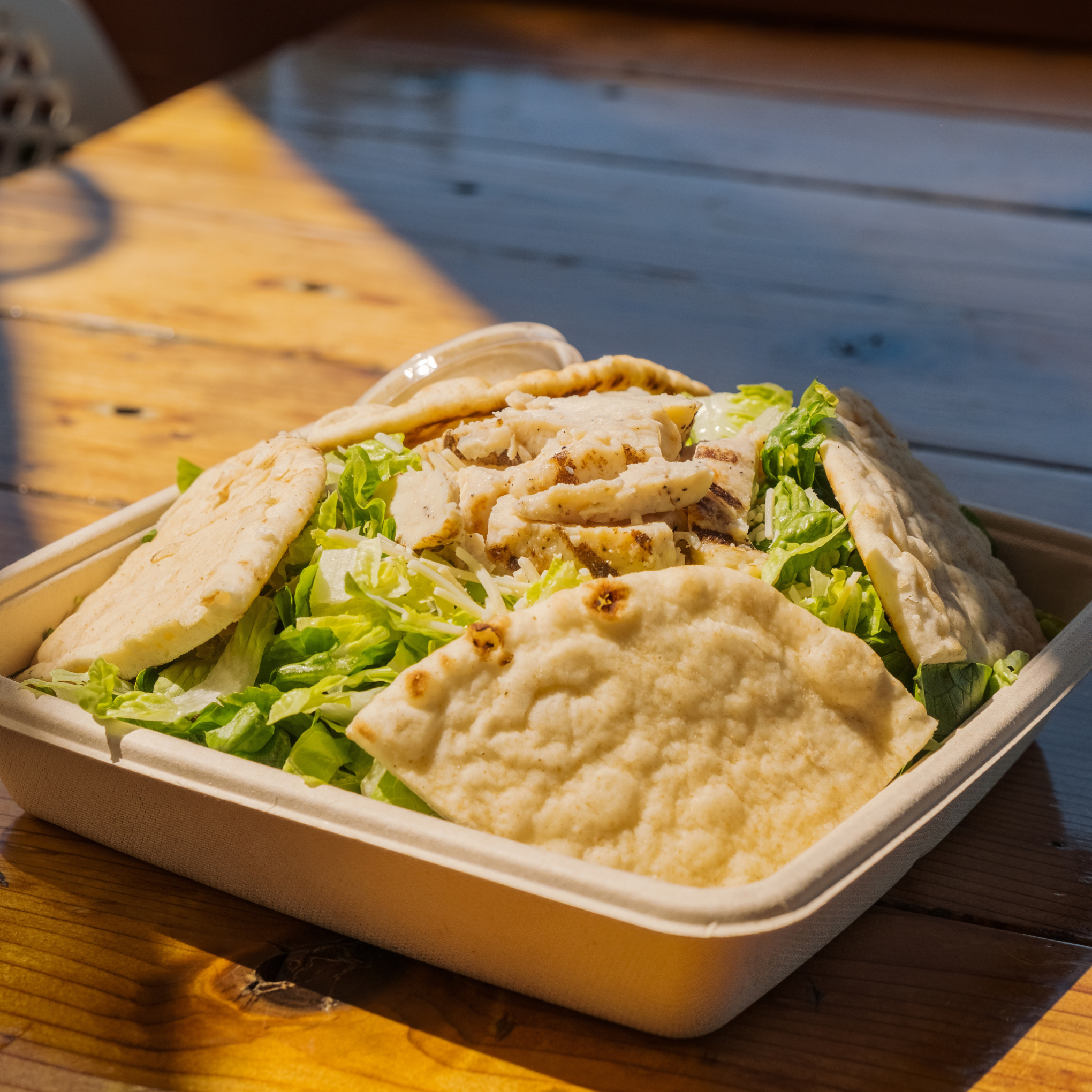 Salad with grilled chicken and flatbread in a takeaway box on a wooden table.