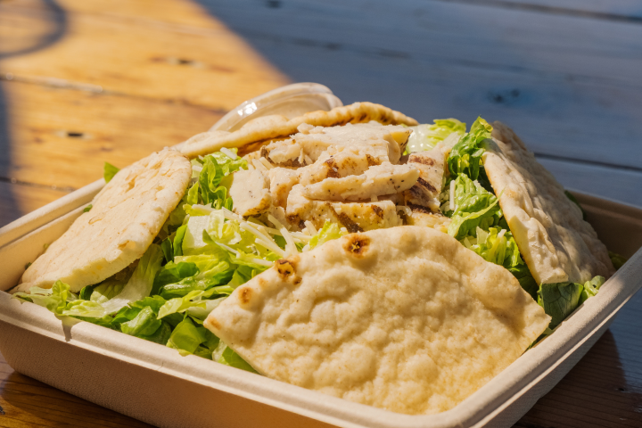 Salad with grilled chicken and flatbread in a takeaway box on a wooden table.