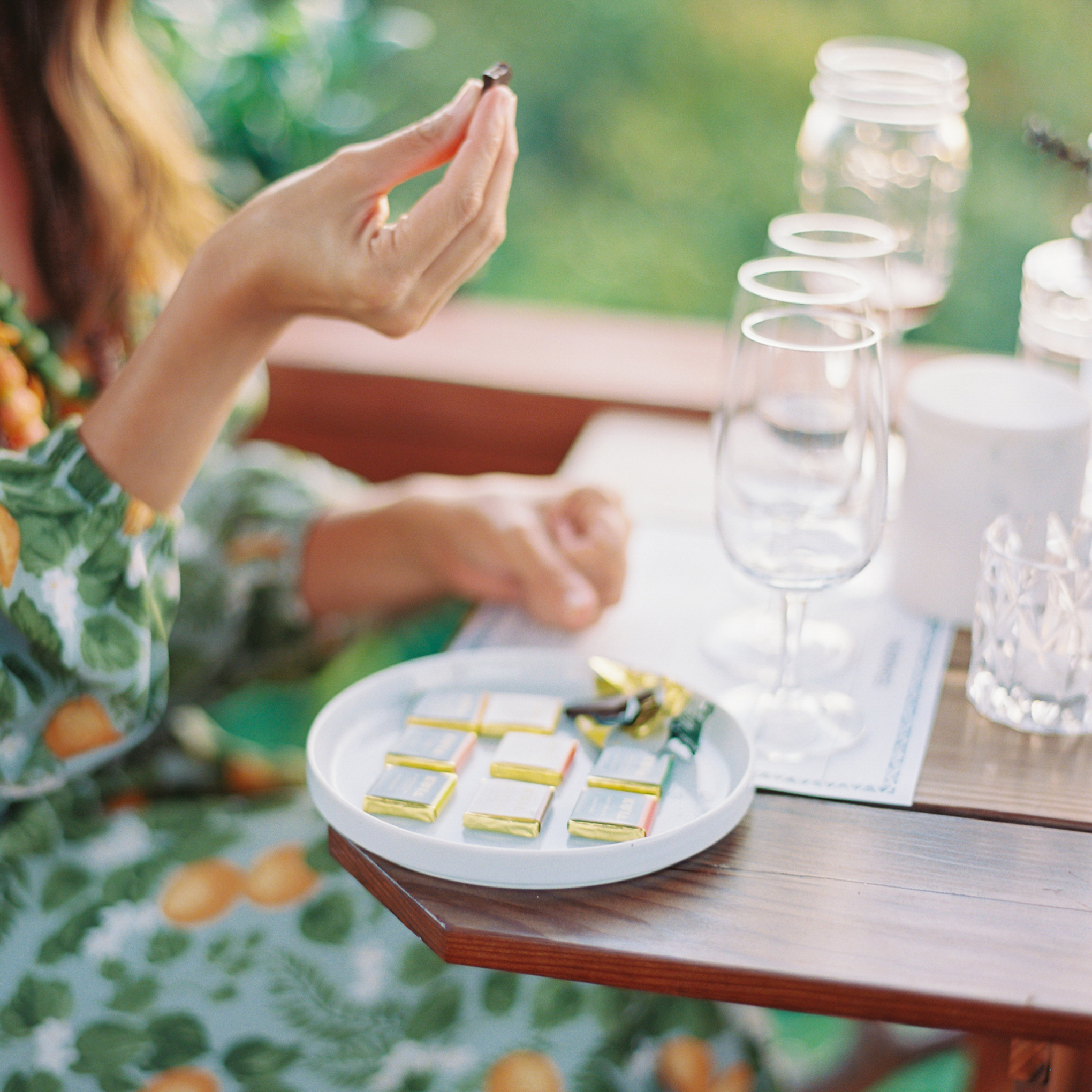 Person holding chocolate piece above plate with wrapped chocolates and wine glasses nearby.