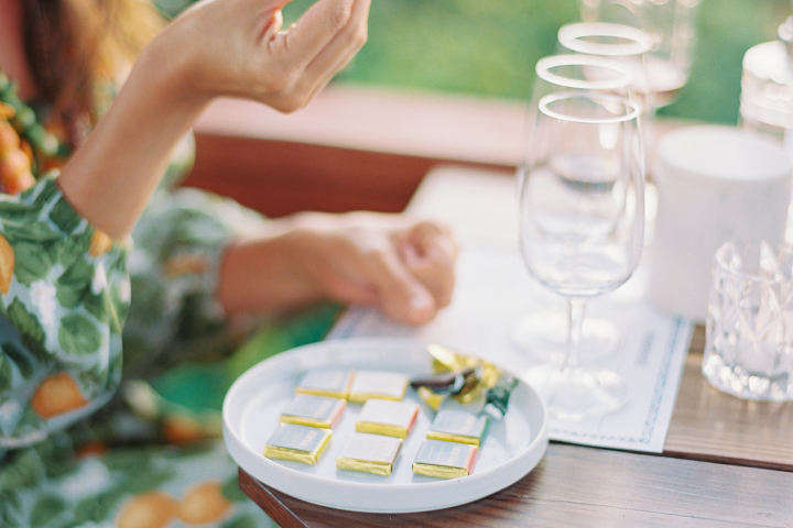 Person holding chocolate piece above plate with wrapped chocolates and wine glasses nearby.