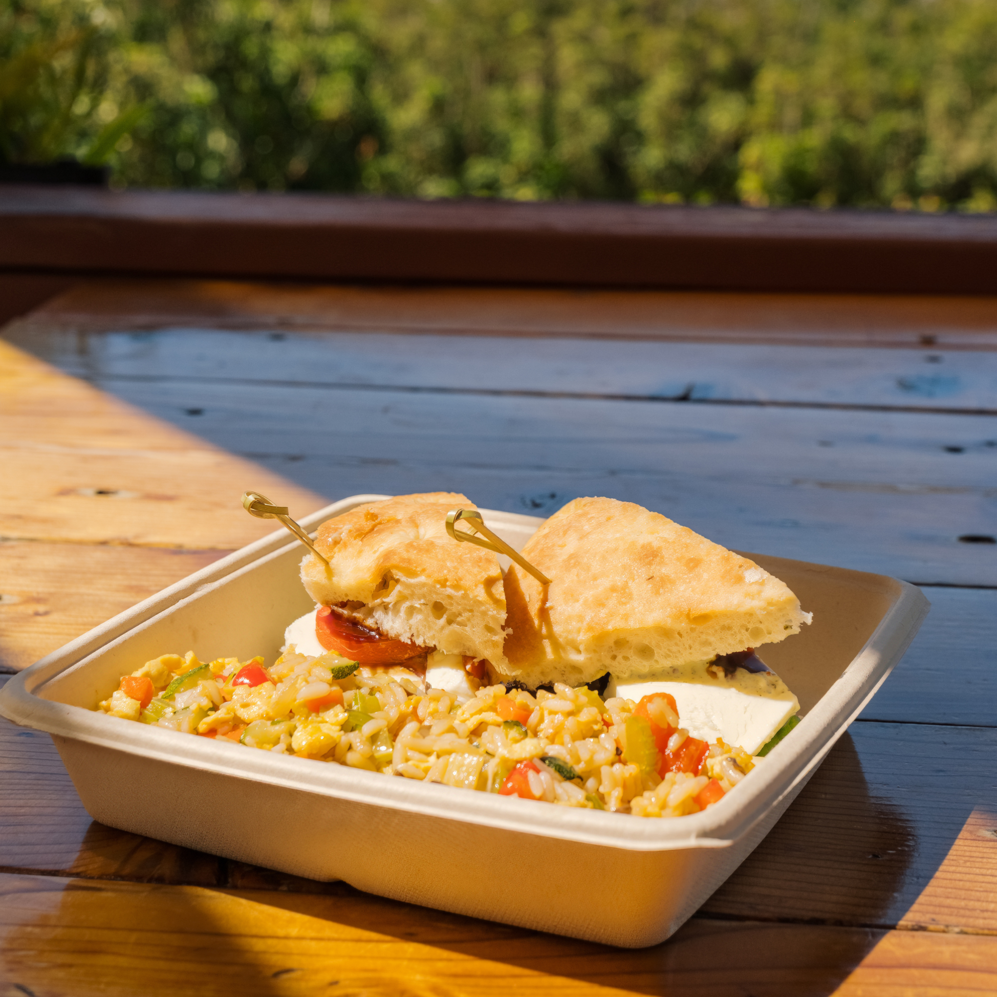 Sandwich and vegetable rice in a tray on a wooden table outdoors.