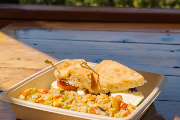 Sandwich and vegetable rice in a tray on a wooden table outdoors.