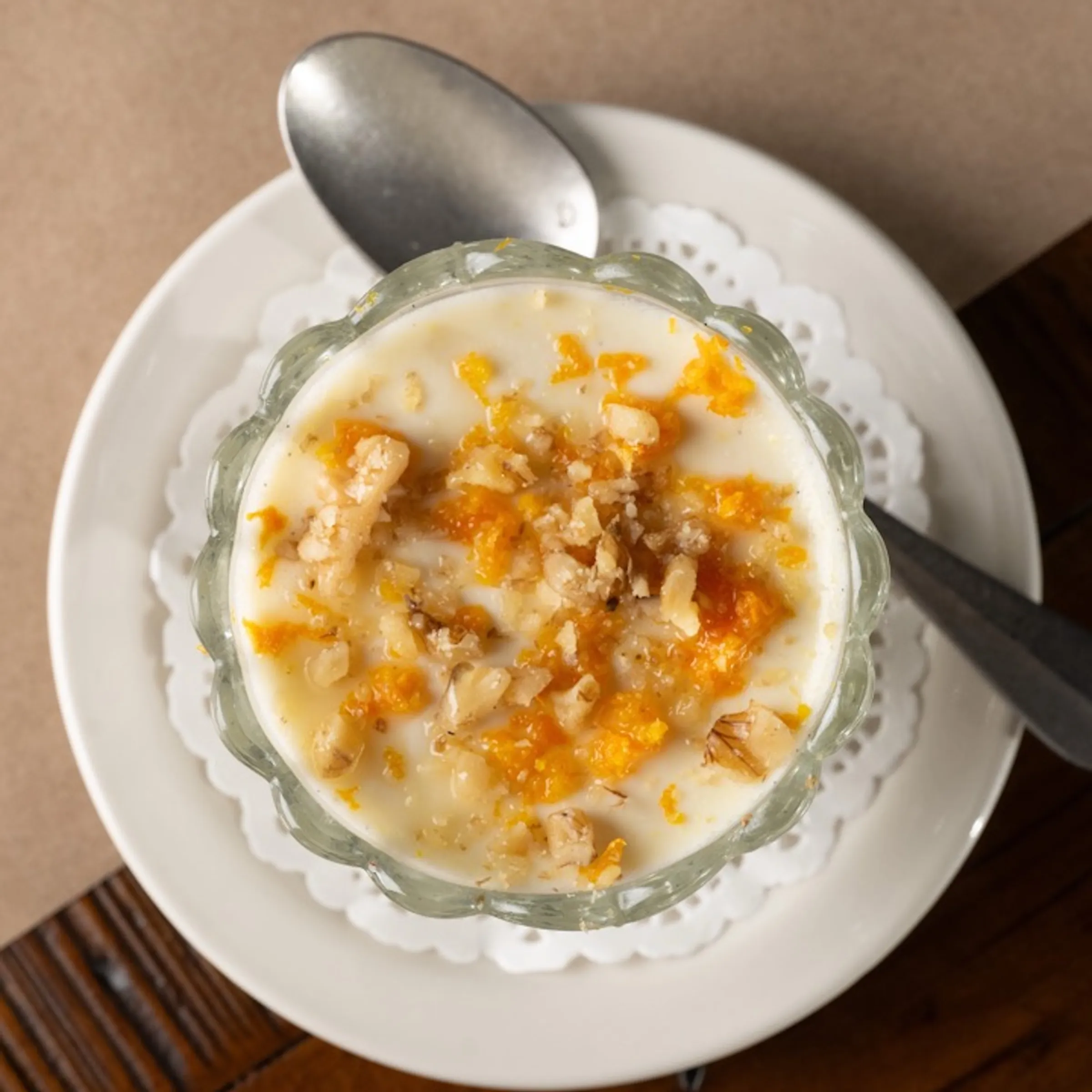Glass bowl of yogurt with orange zest and walnuts on a plate with a spoon.