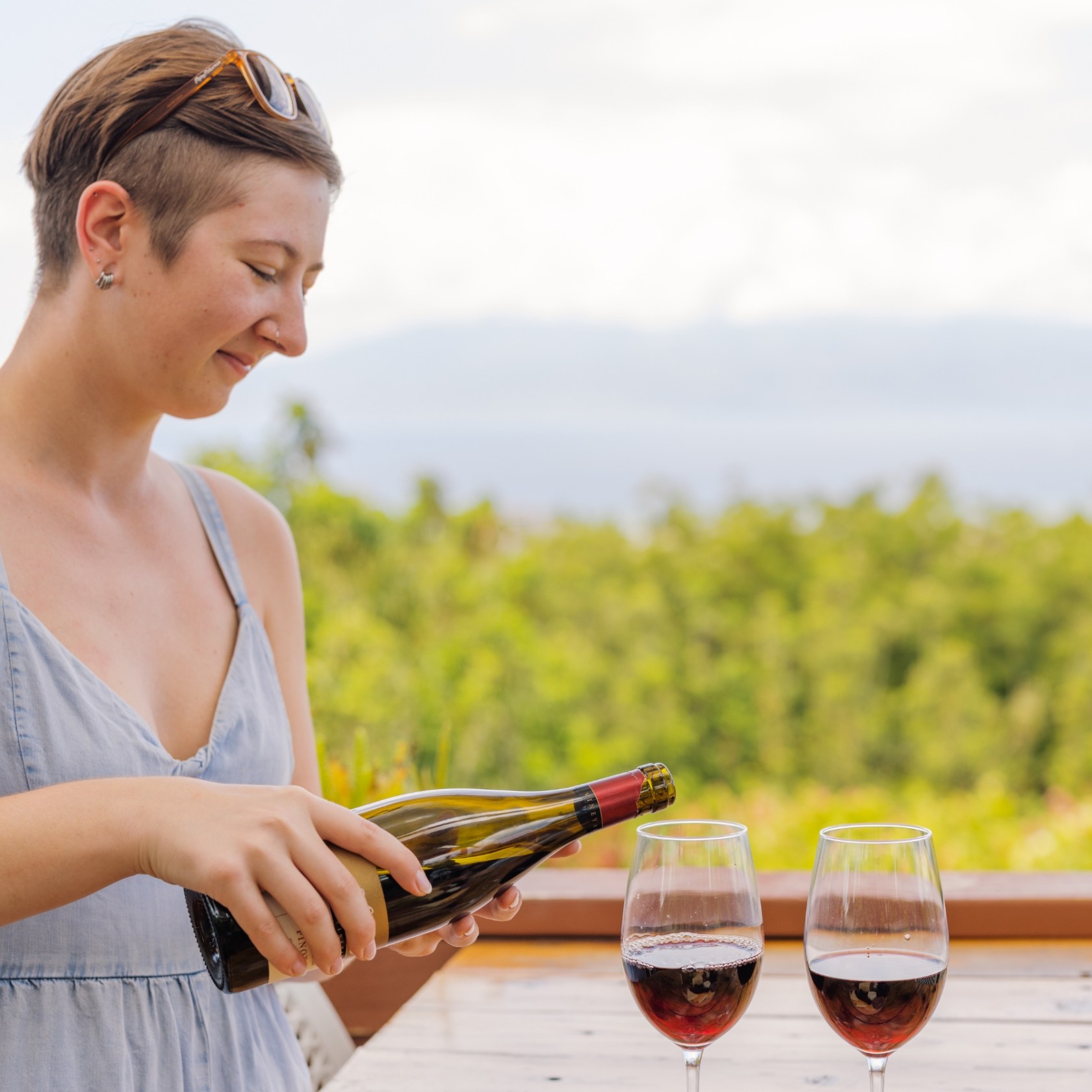 Person smiling, opening a wine bottle outdoors, with two wine glasses on the table.