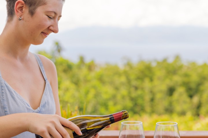 Person smiling, opening a wine bottle outdoors, with two wine glasses on the table.