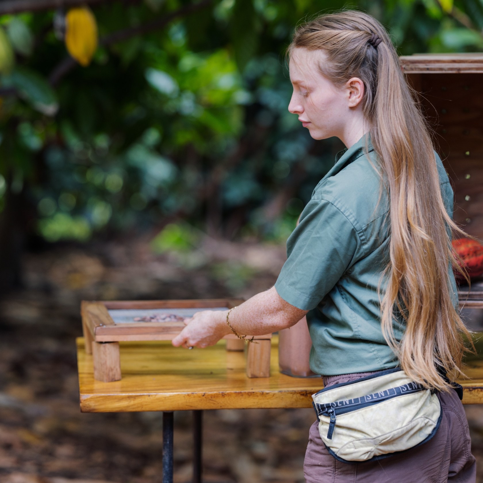 Person with long hair, wearing a green shirt and fanny pack, standing behind a yellow wooden table outside.