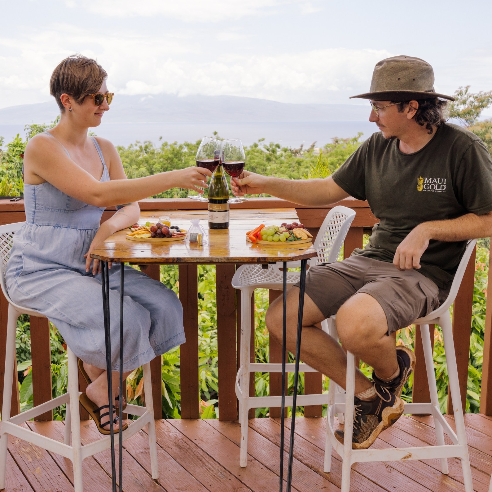 Two people clinking wine glasses at a patio table with food, overlooking a scenic view.