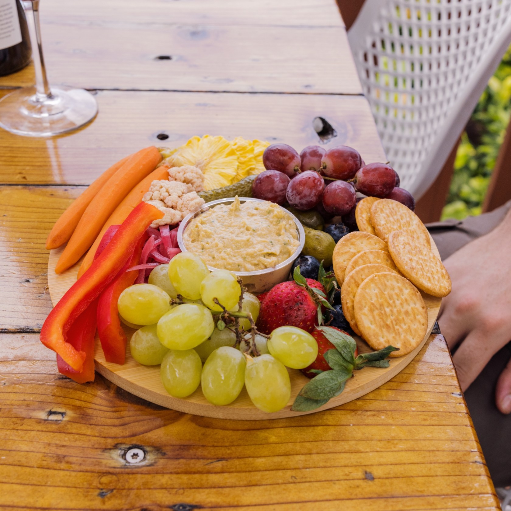 Round wooden platter with grapes, crackers, carrots, hummus, and red pepper strips.