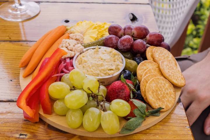 Round wooden platter with grapes, crackers, carrots, hummus, and red pepper strips.